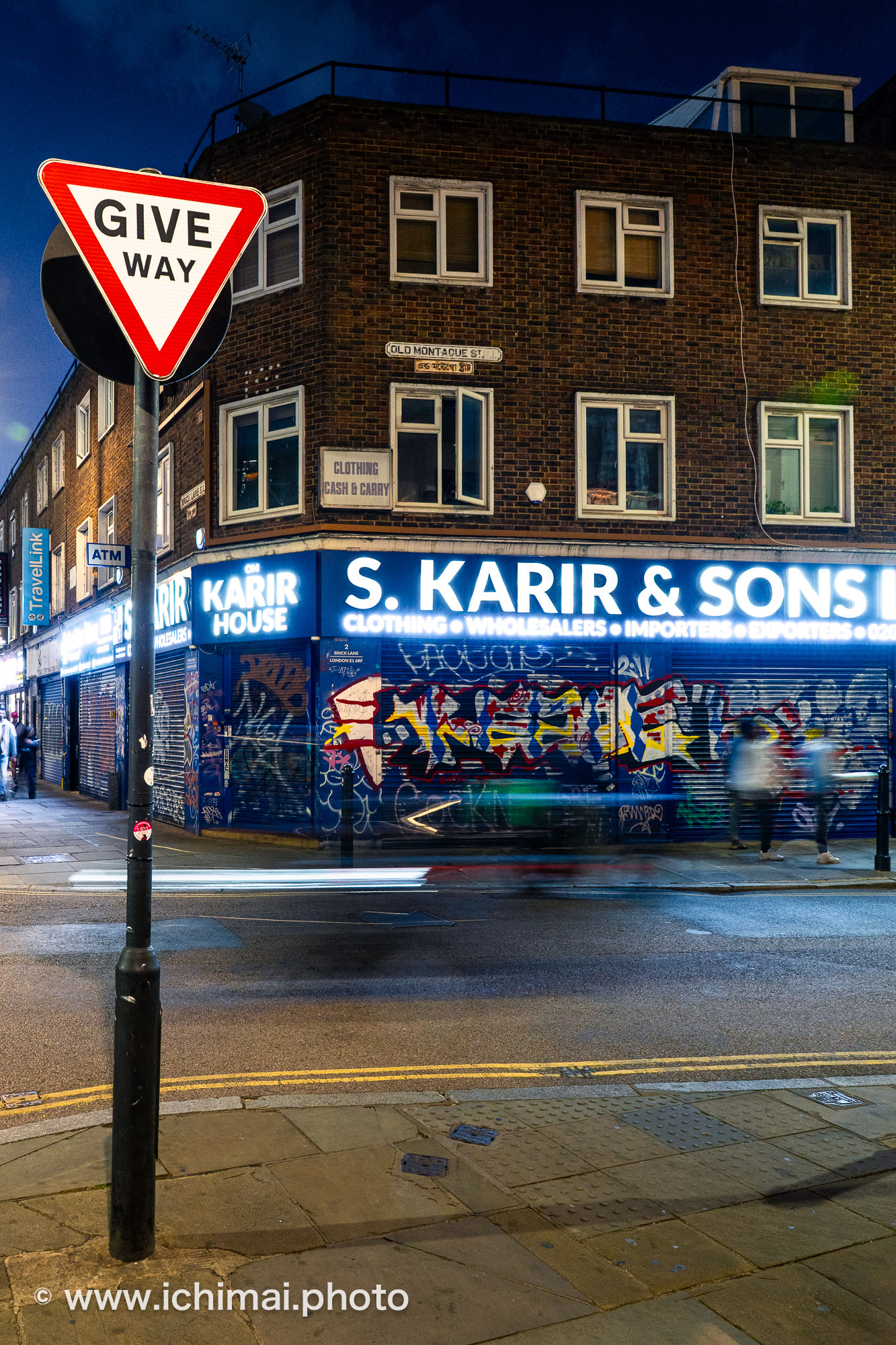 Photographie de rue à Brick Lane, Londres, de nuit : panneau Give Way net au premier plan, passants et phares filés en pose longue devant des rideaux couverts de graffitis