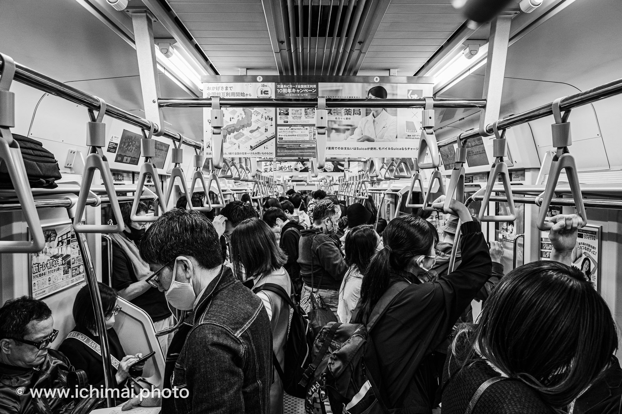 Photographie noir et blanc d’une rame de métro bondée à Tokyo, passagers masqués et têtes baissées, poignées alignées