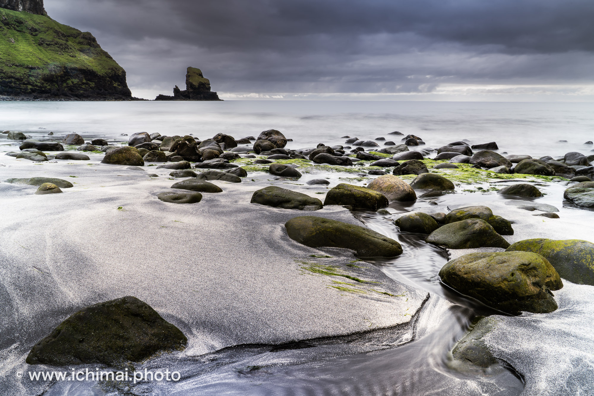 Talisker Bay sur l’île de Skye en Écosse : pose longue d’une plage de sable noir aux rochers moussus, avec un ruisseau sinueux et un piton rocheux sous un ciel chargé