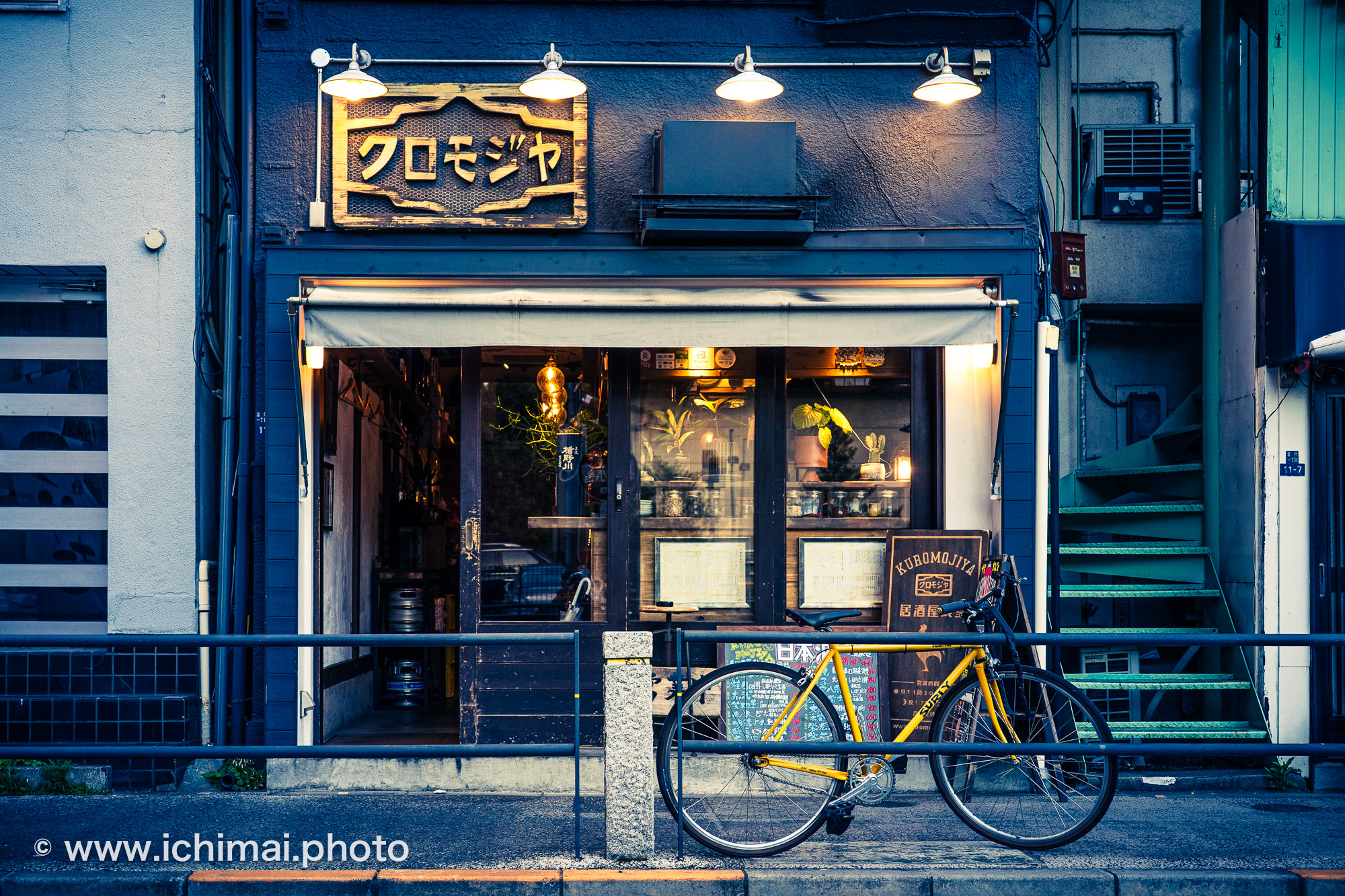 Vélo jaune garé devant la façade bleue d'un izakaya de Tokyo, enseigne en katakana et lampes allumées à la tombée du jour